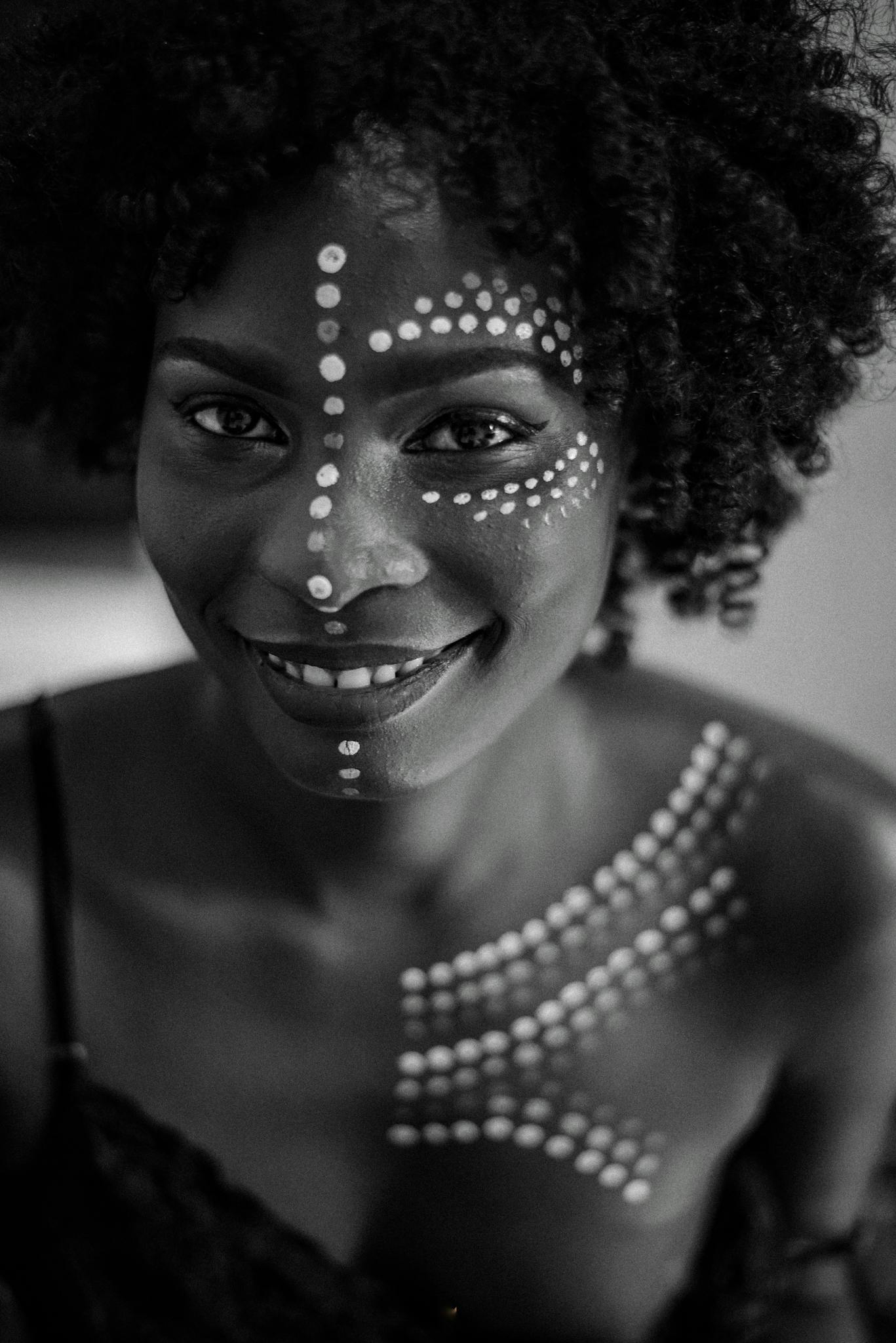 Black and white of cheerful African American female with paint on face and body smiling and looking at camera