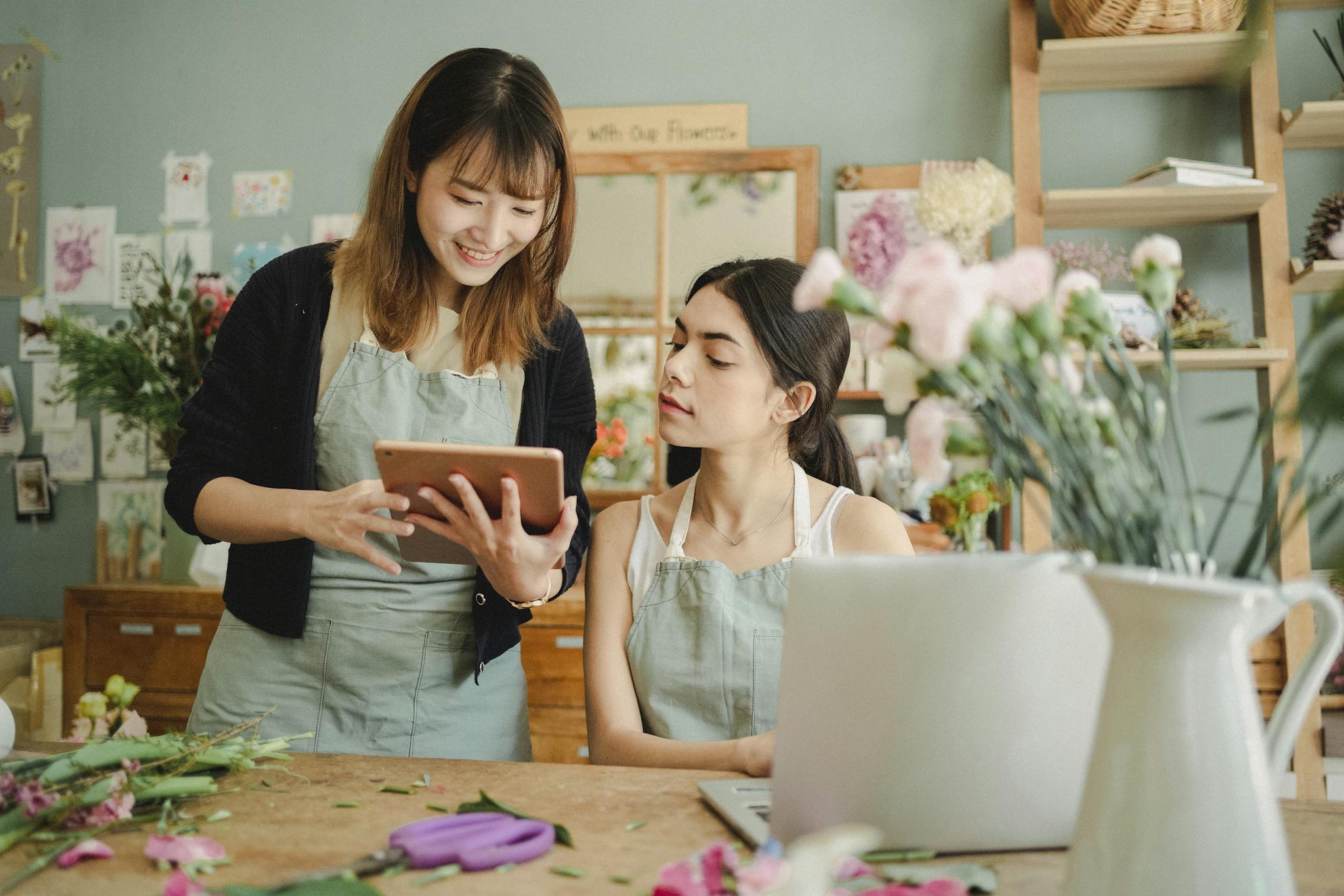 Content multiethnic female coworkers at table with flowers surfing tablet while choosing design for bouquet during work in florist shop