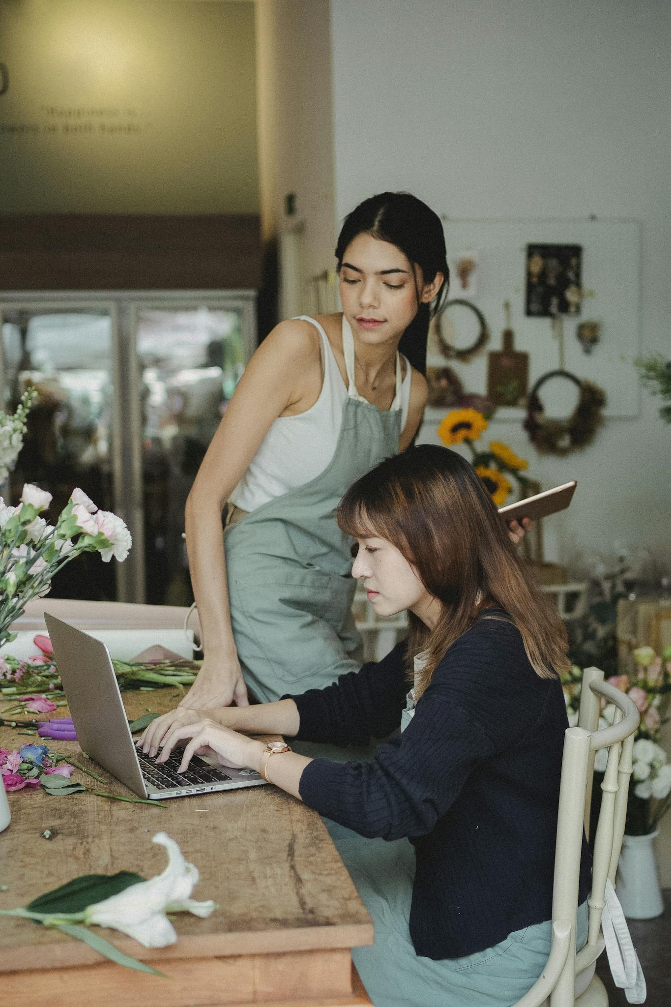 Focused multiethnic female florists sitting at table and typing on computer while checking online orders during work in floral store