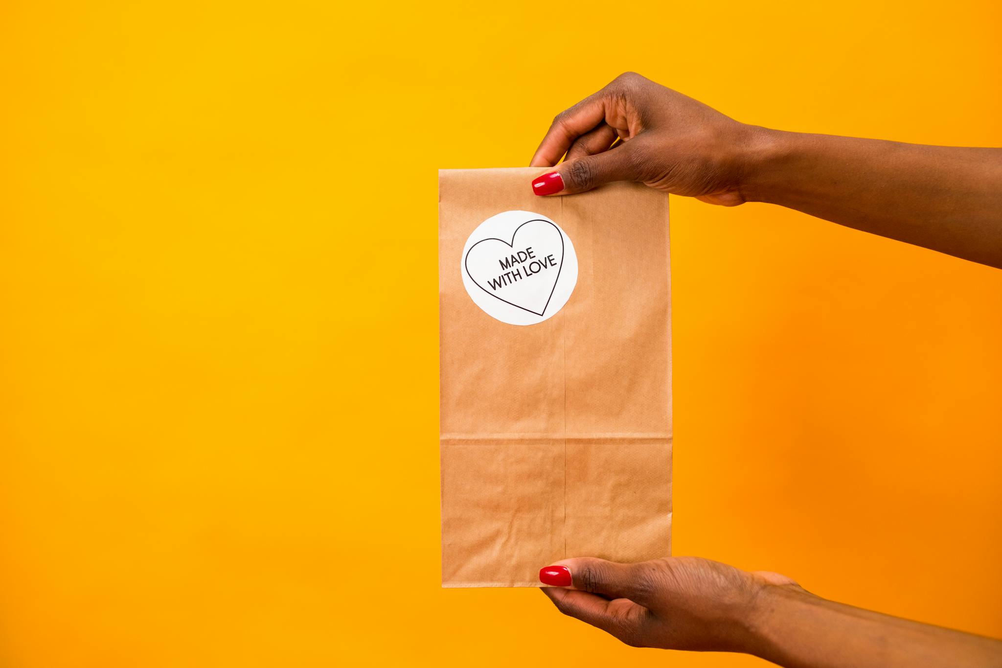 Hands holding a paper bag with 'Made with Love' sticker against a vibrant yellow backdrop.